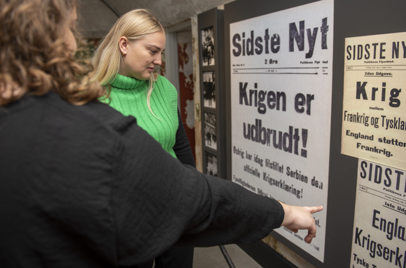 Billedet viser to kvinder, der undersøger en plakat, hvor der står "Krigen er udbrudt!", i udstillingen "På kanten af krig"